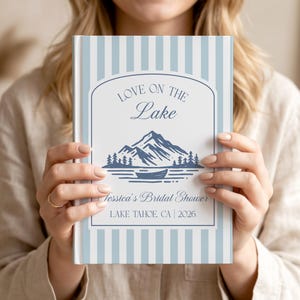 Puede incluir: Un libro de tapa dura con rayas verticales azul claro y blancas. El centro presenta una etiqueta blanca con un gráfico azul oscuro de montañas, bosque y un bote. Texto: LOVE ON THE LAKE, Jessica's Bridal Shower, LAKE TAHOE, CA | 2026.