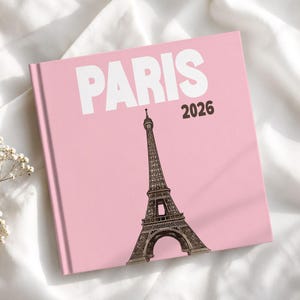 May include: A square book with a light pink cover featuring the word PARIS in large white capital letters and the year 2026 in black text. A detailed black line drawing of the Eiffel Tower is centered below the text on the cover.