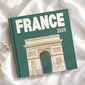 May include: A square hardcover book with a dark green cover featuring the white text FRANCE and the beige text 2026 above a detailed illustration of the Arc de Triomphe in shades of beige, tan, and white with intricate stone carvings.