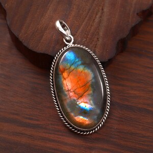 Può includere: Ciondolo ovale in labradorite con un'ansa color argento e un bordo a forma di corda. La pietra preziosa mostra un vivace mix di tonalità arancioni, blu e verdi. Il ciondolo è posto su una superficie di legno scuro.