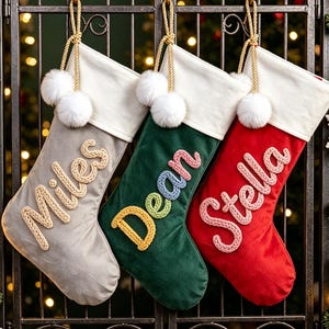Puede incluir: Tres calcetines de Navidad colgados en fila, uno gris con el nombre Miles, uno verde con Dean y uno rojo con Stella. Cada calcetín tiene un puño blanco con dos pompones blancos y cordones dorados para colgar.
