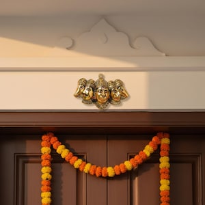 Puede incluir: Un adorno de pared de latón con cinco caras estilizadas, montado sobre una puerta de madera. Las caras tienen tocados ornamentados. Una guirnalda de flores naranjas y amarillas cuelga debajo de la puerta.