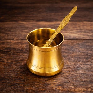 Puede incluir: Una cafetera cilíndrica de color dorado con una cuchara a juego. La cafetera tiene una base ancha y una parte superior más estrecha, con bandas horizontales en el medio. La cuchara descansa dentro de la cafetera, y ambos artículos tienen un acabado brillante.