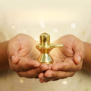 Op de afbeelding: Een goudkleurige religieuze sculptuur met een cilindrische top en een horizontale projectie, vastgehouden in komvormige handen. De sculptuur heeft een uitlopende basis en een glad, gepolijst oppervlak. De achtergrond is een zacht, wazig licht.