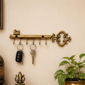 Op de afbeelding: Een wandgemonteerde sleutelhouder in de vorm van een antieke sleutel met een messing afwerking. De horizontale balk is voorzien van zes zilverkleurige metalen haken, met twee sleutelhangers aan de haken, één met een zwarte autosleutel en een metalen sleutel.