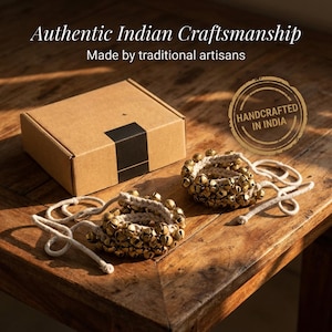 May include: Two sets of brass bells on a cream-colored woven base, with cream-colored cords, sit on a wooden surface. A cardboard box with a black stripe is in the background. Text reads "Authentic Indian Craftsmanship" and "Handcrafted in India."