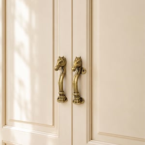 Puede incluir: Dos manijas de puerta de latón antiguo con forma de cabezas y cuellos de caballo, montadas verticalmente en un par de puertas de armario de color crema. Los herrajes de metal presentan tallas detalladas de la crin, los ojos y el hocico.