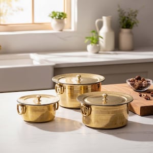 May include: Three gold-coloured metal cooking pots with lids and handles are arranged on a white surface. The pots vary in size, with a small, medium, and large pot. The lids have a small knob on top. The pots are in a kitchen setting.