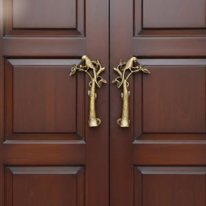 以下が含まれることがあります： 装飾された真鍮製のドアノブのペア。各ノブは、木の枝に止まっている鳥が特徴です。ノブは、パネルデザインの濃い茶色の木製のドアに取り付けられています。ノブの高さは約15cmです。