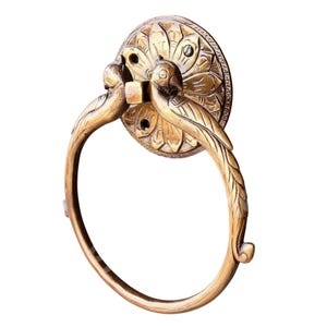 May include: A brass towel ring with a decorative floral backplate. Two bird figures are perched on the ring, which is attached to the backplate. The ring is a warm gold color.