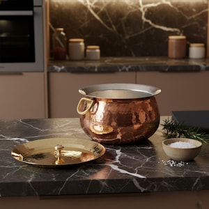 Puede incluir: Una olla de cocina de cobre con acabado martillado y asas de latón, junto con una tapa dorada. La olla está sobre una encimera de mármol negro, con un pequeño cuenco de sal y romero. Utensilios de cocina.