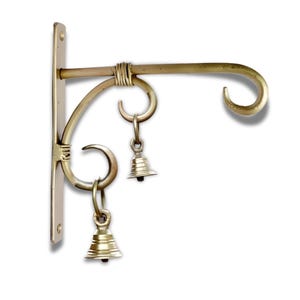 May include: A brass-coloured wall-mounted plant hanger with a curved hook and decorative scrollwork. Two small bells hang from the scrollwork. The hanger is designed to hold a hanging basket or other decorative items.