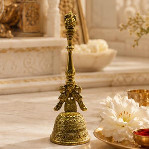 May include: A tall, metallic gold-toned hand bell featuring an intricate embossed pattern on the base and a handle topped with a small, detailed figure of Ganesha. The bell has a flared bottom and a slender, decorative stem with peacock motifs.