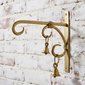 Puede incluir: Un soporte de metal dorado para colgar con un brazo curvo y dos pequeñas campanas. El soporte está montado en una pared de ladrillo blanco. El diseño presenta remolinos decorativos y una sección enrollada.