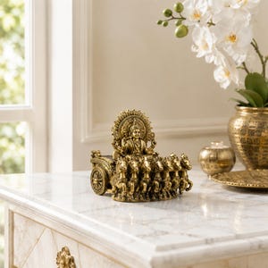 May include: A gold-toned metallic figurine depicting a deity seated in a chariot pulled by seven horses. The figure features an ornate circular halo behind the head, with intricate detailing on the chariot wheels and the horses' harnesses.
