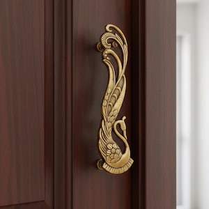 Op de afbeelding: Decoratieve messing deurklink met een pauwontwerp. De klink is gemonteerd op een donkerbruine houten deur. Het lichaam van de pauw is gedetailleerd met veren en bloemmotieven, en de klink buigt sierlijk.