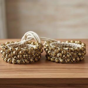 May include: Two sets of ankle bells. Each set features rows of small, gold-colored bells attached to a cream-colored woven band. A cream-colored cord connects the two sets. The bells are arranged on a wooden surface.