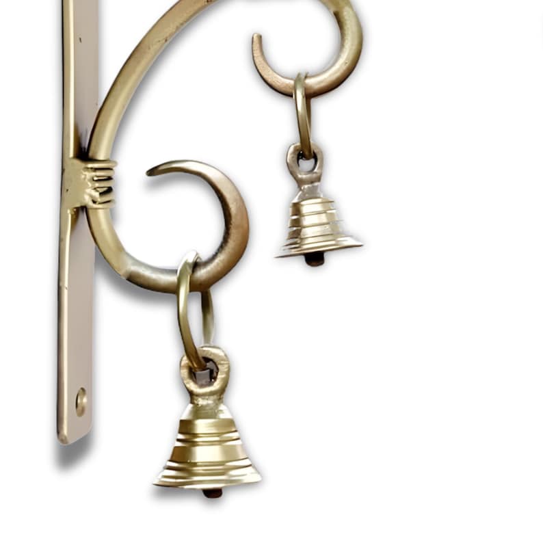 May include: A brass-coloured wall-mounted bracket with a decorative scroll design. Two small, bell-shaped ornaments hang from the bracket. The bracket is designed to hold a hanging item, such as a wind chime or plant.