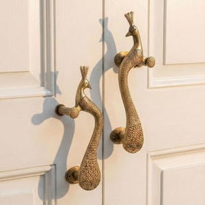 May include: Two brass peacock-shaped door handles. The handles are detailed with feathers and stand out against a white door. The handles are approximately 10 inches long.
