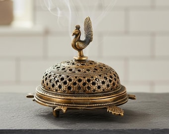 May include: A brass incense burner with a peacock finial. The domed lid features intricate cut-out patterns, and the base has a turtle-like design. Wisps of smoke rise from the burner, suggesting its use. The burner is on a dark gray surface.