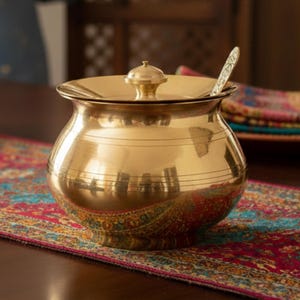 Op de afbeelding: Een goudkleurige, ronde serveerschaal met deksel en een kleine knop bovenop. De kom heeft horizontale geëtste lijnen en een kleine lepel op de rand. Hij staat op een kleurrijke patroonloper, wat wijst op een keuken of eetkamer.