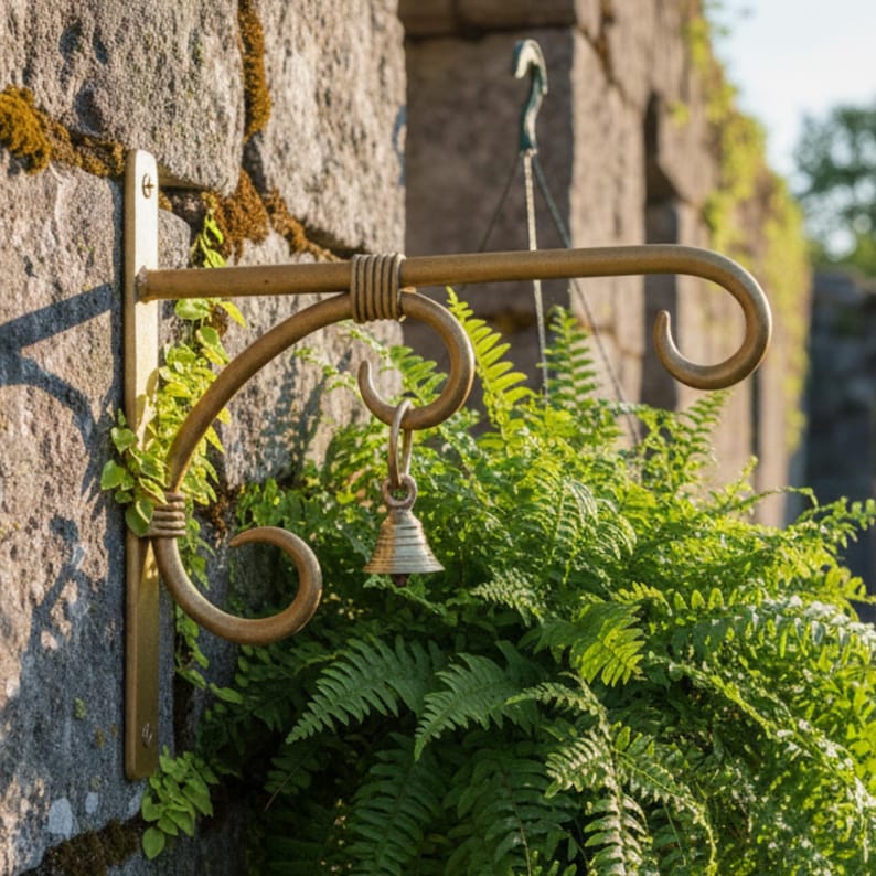 May include: A decorative gold-coloured metal plant hanger fixed to a stone wall. The hanger has a curved design with a small bell suspended from it. Green ferns are below the hanger, creating a natural contrast. The wall is made of stone.