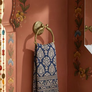 May include: A brass towel ring with a decorative floral design holds a blue and white patterned towel. The ring is mounted on a reddish-brown wall with floral wall art. The towel has a detailed, symmetrical pattern.