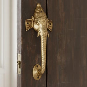 May include: A brass door handle in the shape of an elephant's head and trunk, mounted on a dark wooden door. The handle features intricate details and a decorative design, adding a unique touch to the door's appearance.