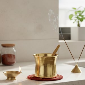 May include: A brass incense burner with a wooden spoon inside, sitting on a red coaster with gold detailing. A small brass oil lamp with a lit flame is to the left. Incense sticks are burning, emitting smoke.