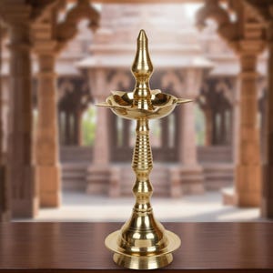 Może przedstawiać: Wysoka, ozdobna lampa mosiężna z gwiaździstym uchwytem na olej i dekoracyjnym, żebrowanym trzonem. Lampa ma rozszerzoną podstawę i spiczasty wierzchołek. Tło jest rozmyte, pokazujące budynek z kolumnami.