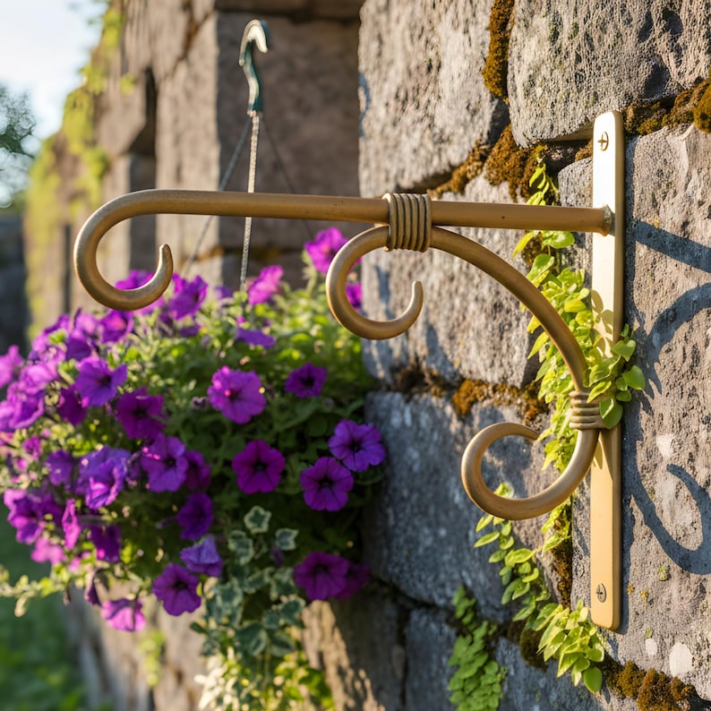 May include: A decorative gold-coloured metal hanging basket bracket fixed to a stone wall. The bracket features a curved design with scrolled details, supporting a hanging basket overflowing with purple petunias. Greenery adds a natural accent.