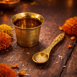 Puede incluir: Una taza y una cuchara de latón dorado descansan sobre una superficie de madera, con arroz en la cuchara. La cuchara tiene un mango adornado. Flores naranjas y amarillas y velas encendidas en el fondo.