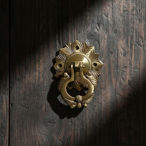 May include: A brass door knocker with a floral design. The knocker is mounted on a dark wooden door. The knocker has a circular centre with a ring handle and is surrounded by a decorative, petal-like pattern.