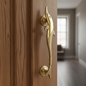 Könnte beinhalten: Ein messingfarbener Türgriff in Form eines Delfins ist an einer Holztür befestigt. Der Griff weist ein detailliertes Design eines Delfins mit strukturiertem Körper und Flossen auf. Die Tür hat eine warme braune Farbe, und der Hintergrund ist unscharf.