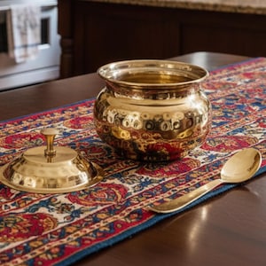 Puede incluir: Un cuenco dorado de metal martillado con tapa y cuchara, sobre un corredor estampado. El cuenco tiene forma redondeada y tapa con asa pequeña. La cuchara es larga y delgada. El corredor presenta un diseño floral rojo, azul y blanco.