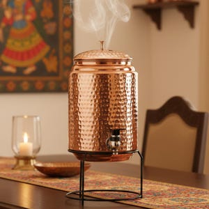 May include: A copper water dispenser with a hammered texture, a lid, and a spigot. It sits on a black metal stand. Steam rises from the top. A candle in a glass holder and a decorative table runner are in the background.