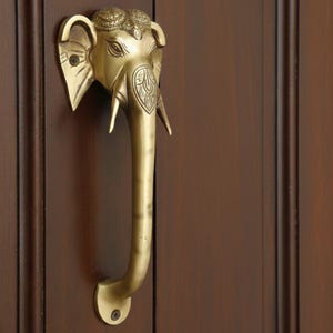 Peut inclure: Une poignée de porte en laiton en forme d'éléphant. La poignée est ornée d'une tête, d'une trompe et de défenses d'éléphant. La poignée est fixée sur une porte en bois brun foncé. La tête d'éléphant présente des sculptures et des détails complexes.