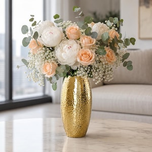 Könnte beinhalten: Eine hohe, gehämmert goldfarbene Metallvase enthält ein großes, üppiges Blumenarrangement. Der Strauß besteht aus weißen Pfingstrosen, zarten pfirsichfarbenen Rosen, feinem weißem Schleierkraut und runden grünen Eukalyptusblättern in runder Form.