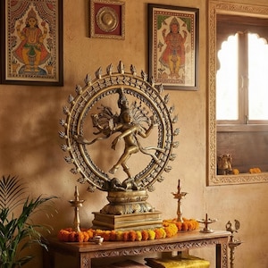 May include: A bronze statue of a dancing deity, framed by a circular halo, sits on a wooden table decorated with marigold garlands. Framed artwork and decorative lamps enhance the scene. The room has a warm, inviting atmosphere. The statue is the focal point.