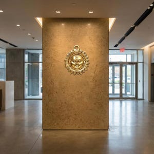 Könnte beinhalten: Ein goldfarbener, sonnenförmiger Wandschmuck mit einem lächelnden Gesicht in der Mitte. Die Sonne hat detaillierte Strahlen und eine Schlaufe zum Aufhängen. Die Wand hat eine strukturierte, braune Oberfläche.