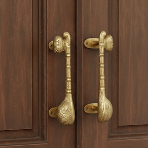 Op de afbeelding: Twee sierlijke messing deurklinken met ingewikkelde bloemen- en geometrische patronen. De klinken zijn gemonteerd op een donkerbruine houten deur, wat een vintage of antieke uitstraling geeft. De klinken hebben een unieke, langwerpige vorm.