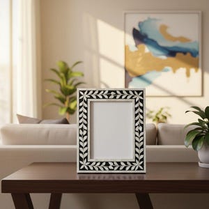 Puede incluir: Un marco de fotos rectangular con un diseño floral en blanco y negro. El marco tiene un interior blanco y está hecho de un material oscuro. El patrón floral presenta hojas blancas y detalles en negro. El marco se muestra sobre una mesa marrón oscuro.