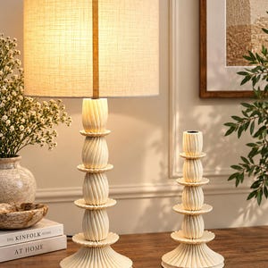 May include: A table lamp and a matching candle holder, both featuring a cream-colored, stacked, spiraled design with a textured finish. The lamp has a cylindrical, light-colored fabric shade that emits a warm glow from the internal light source.
