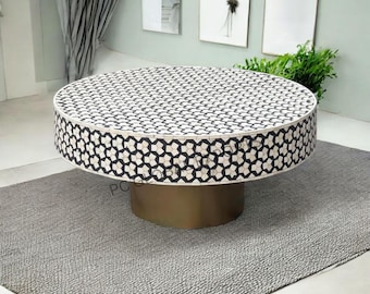 Op de afbeelding: Een ronde salontafel met een zwart-wit geometrisch sterpatroon ingelegd op de bovenkant en de zijkanten. De tafel wordt ondersteund door een stevige, cilindrische voet met een metallic messing afwerking, wat zorgt voor een stabiele basis.