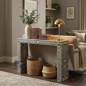 Può includere: Un tavolo console rettangolare con un motivo floreale a intarsio in osso bianco e nero. Il tavolo sostiene un alto vaso in ceramica con rami di eucalipto, una pila di cinque libri con copertina rigida e una lampada in metallo dorato a cupola.