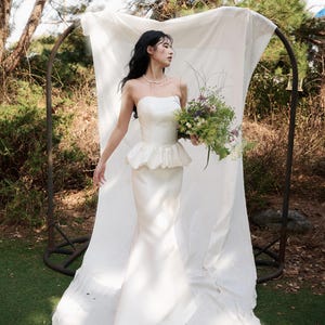 May include: A strapless ivory wedding gown with a peplum waist and a mermaid silhouette. The bride holds a bouquet of flowers. The background includes a white fabric backdrop and greenery.
