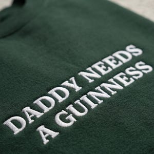 May include: Dark green sweatshirt with the embroidered text "DADDY NEEDS A GUINNESS" in white. The text is in a bold, sans-serif font. The sweatshirt appears to be made of a soft, textured fabric.