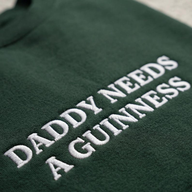 May include: Dark green sweatshirt with the embroidered text "DADDY NEEDS A GUINNESS" in white. The text is in a bold, sans-serif font. The sweatshirt appears to be made of a soft, textured fabric.