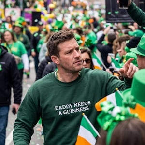 May include: A green sweatshirt with the text "DADDY NEEDS A GUINNESS" in white. The image shows a man in a crowd, likely at a St. Patrick's Day parade, with people wearing green and holding Irish flags.