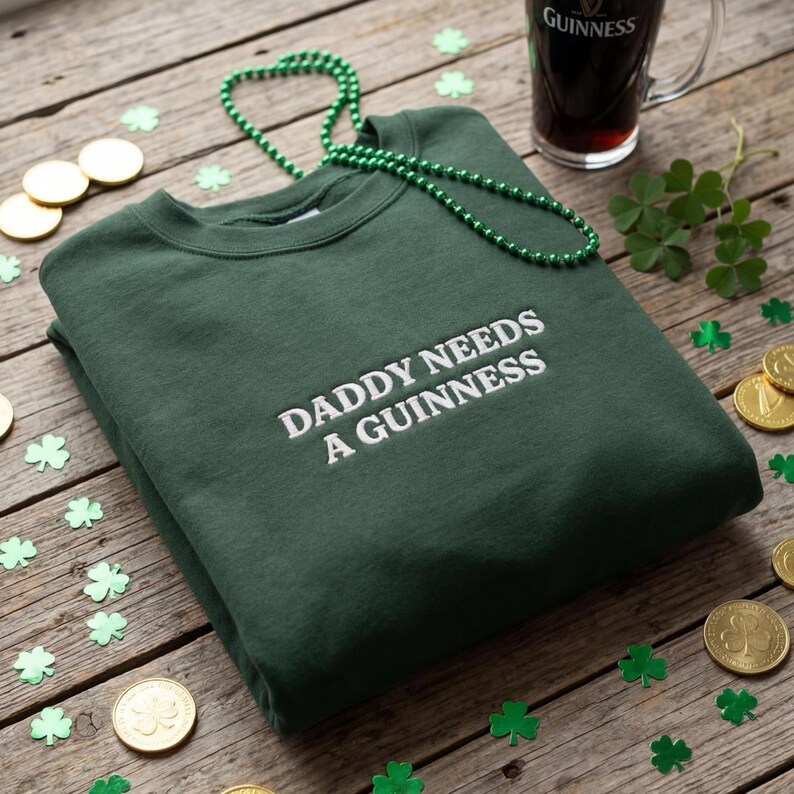 May include: A dark green sweatshirt with the embroidered text "DADDY NEEDS A GUINNESS". The image includes a green beaded necklace, gold coins, shamrock confetti, and a pint of Guinness.
